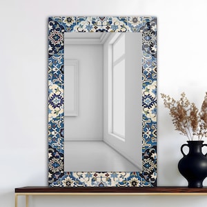 K&ouml;nnte beinhalten: Rechteckiger Spiegel mit dekorativem Rand mit blauem, wei&szlig;em und beigem Blumenmuster. Der Spiegel steht auf einem dunklen Holzregal, rechts daneben eine schwarze Vase und getrocknete Pflanzen.