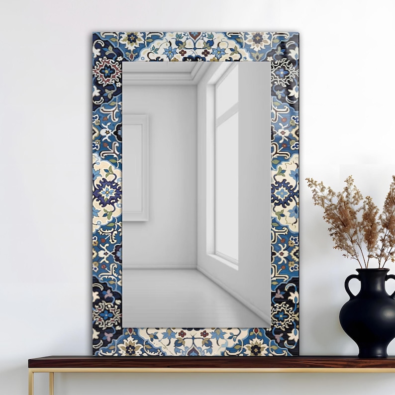 K&ouml;nnte beinhalten: Rechteckiger Spiegel mit dekorativem Rand mit blauem, wei&szlig;em und beigem Blumenmuster. Der Spiegel steht auf einem dunklen Holzregal, rechts daneben eine schwarze Vase und getrocknete Pflanzen.