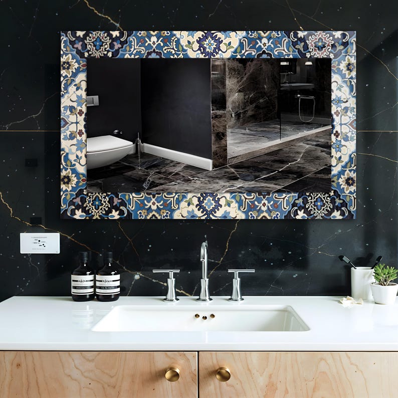 K&ouml;nnte beinhalten: Rechteckiger Spiegel mit dekorativem Rand mit blauen, wei&szlig;en und beigen Blumenmustern. Der Spiegel ist an einer schwarzen Marmorwand &uuml;ber einem wei&szlig;en Waschbecken und einem Holzschrank mit Messingkn&ouml;pfen angebracht. Die Spiegelung zeigt ein Badezimmer.