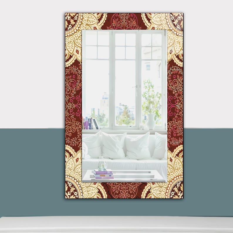 May include: Rectangular mirror with a decorative border featuring a repeating paisley pattern in burgundy, cream, and pink. The mirror reflects a room with a white sofa, pillows, and a window with a view.