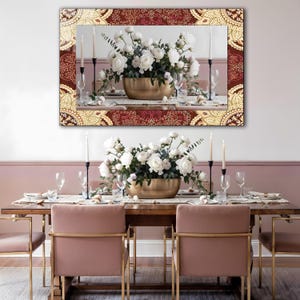 May include: A dining room scene featuring a large mirror with a decorative burgundy and gold frame above a wooden table set for a meal. The table is adorned with a floral centerpiece, candles, and place settings. Pink chairs with gold frames complete the elegant setting.