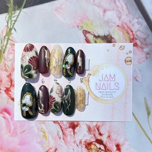May include: A set of press-on nails with various designs. Some nails feature floral patterns in red, green, and white. Others have gold leaf accents, chains, and gemstones. The nails are displayed on a card with the text "JAM NAILS".