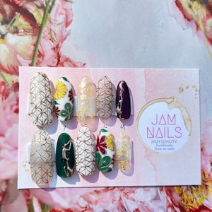 May include: A set of press-on nails with various designs. Some nails feature gold geometric patterns, floral designs with red and yellow accents, and gold leaf details. The nails are displayed on a pink card with the text "JAM NAILS".