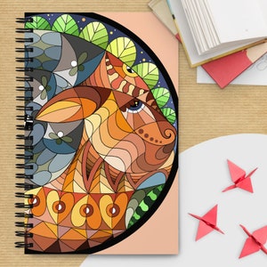 May include: Spiral-bound notebook featuring a colorful, geometric design of an animal's face. The artwork incorporates shades of brown, orange, blue, and green. The notebook is open, revealing blank pages, with origami cranes nearby.