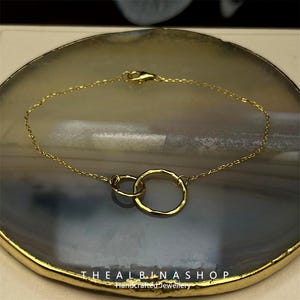 May include: A gold chain bracelet with two interlocked circles. The circles are hammered and have a textured finish. The bracelet is on a gray and white background.