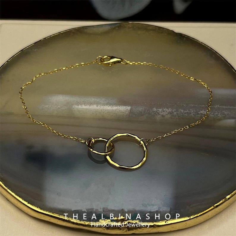 May include: A gold chain bracelet with two interlocked circles. The circles are hammered and have a textured finish. The bracelet is on a gray and white background.