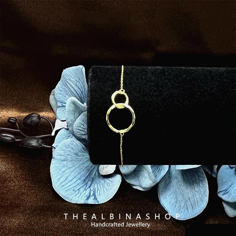 May include: A delicate gold chain bracelet with two interlocking circles. The circles are textured and have a slightly hammered finish. The bracelet is on a black background.