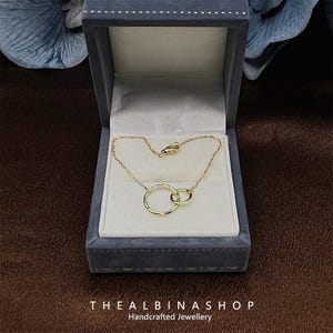 May include: A gold necklace with two interlocking circles, displayed in a gray velvet box. The box has the text "THE ALBINASHOP Handcrafted Jewellery" on the bottom.