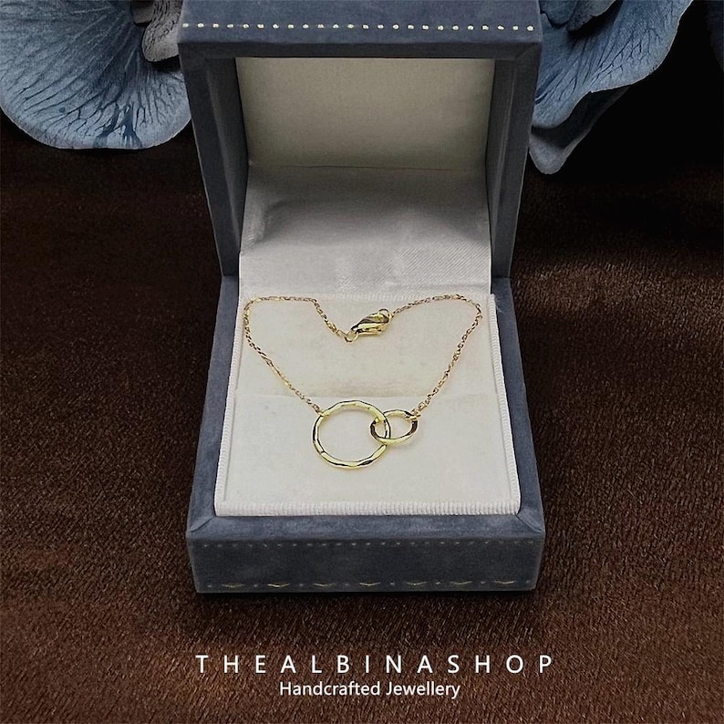 May include: A gold necklace with two interlocking circles, displayed in a gray velvet box. The box has the text "THE ALBINASHOP Handcrafted Jewellery" on the bottom.
