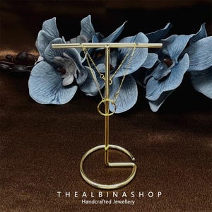 May include: Two gold chains with a gold ring pendant displayed on a gold metal stand. The stand has a round base and a T-shaped top. The stand is on a brown surface.