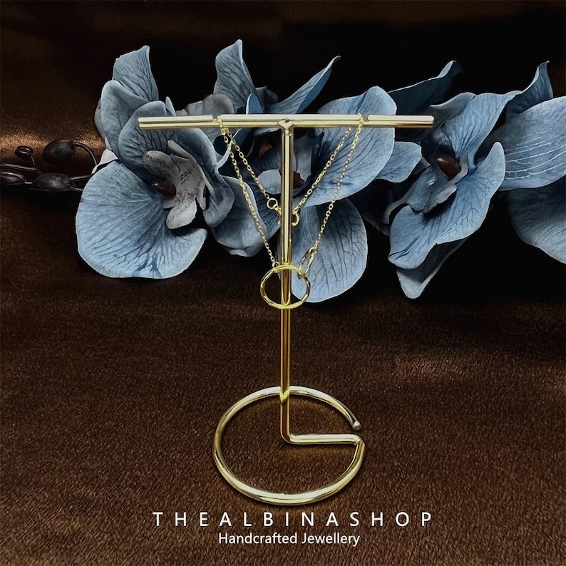 May include: Two gold chains with a gold ring pendant displayed on a gold metal stand. The stand has a round base and a T-shaped top. The stand is on a brown surface.