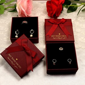 Puede incluir: Tres cajas de regalo de terciopelo rojo con letras doradas que dicen "Mejores deseos para ti, especialmente para ti". Las cajas est&aacute;n decoradas con lazos de sat&eacute;n rojo y contienen joyas, incluyendo un anillo, aretes y un colgante.