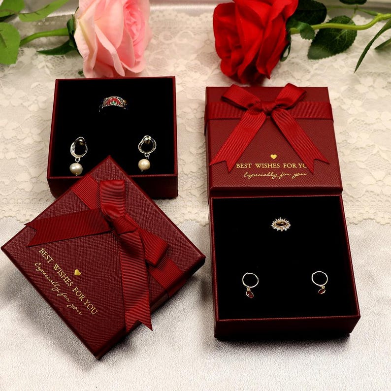 Puede incluir: Tres cajas de regalo de terciopelo rojo con letras doradas que dicen "Mejores deseos para ti, especialmente para ti". Las cajas est&aacute;n decoradas con lazos de sat&eacute;n rojo y contienen joyas, incluyendo un anillo, aretes y un colgante.