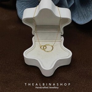 May include: A gold necklace with a delicate chain and two interlocking circles pendant. The necklace is displayed in a white star-shaped box. The text "THEALBINASHOP Handcrafted Jewellery" is visible at the bottom of the image.