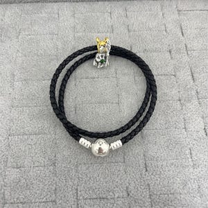 May include: A black braided leather wrap bracelet featuring a silver-toned spherical clasp and a metallic charm depicting a figure with gold-colored horns, a silver body, and a small green accent detail positioned at the top of the circular cord.
