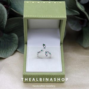 Könnte beinhalten: Ein silberner Schlangenring mit grünen Edelsteinen, präsentiert in einer grünen Samtbox. Die Box trägt den Text "Thealbinashop" und "Handgefertigter Schmuck".