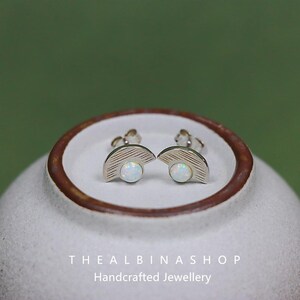 Puede incluir: Un par de pendientes de plata con un dise&ntilde;o de medio c&iacute;rculo y una piedra de &oacute;palo blanco en el centro. Los pendientes est&aacute;n sobre una superficie de cer&aacute;mica blanca con el texto "The Albina Shop Handcrafted Jewellery".