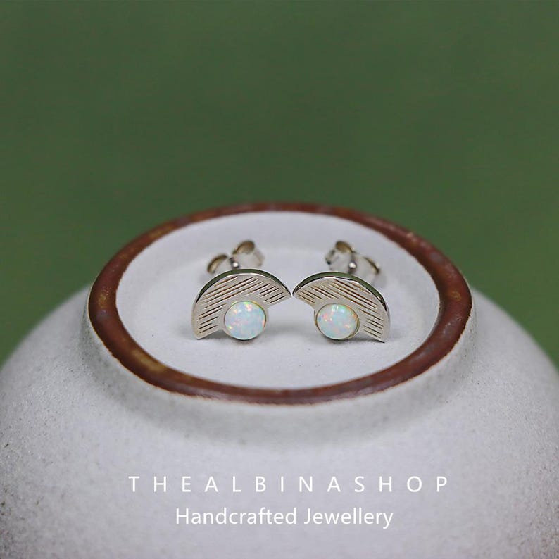 Puede incluir: Un par de pendientes de plata con un dise&ntilde;o de medio c&iacute;rculo y una piedra de &oacute;palo blanco en el centro. Los pendientes est&aacute;n sobre una superficie de cer&aacute;mica blanca con el texto "The Albina Shop Handcrafted Jewellery".