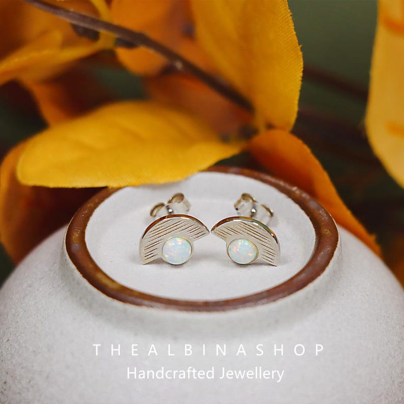 Puede incluir: Un par de pendientes de plata con forma de media luna y una piedra de &oacute;palo blanco en el centro. Los pendientes se muestran en un plato de cer&aacute;mica blanca con un borde marr&oacute;n. El texto "The Albina Shop Handcrafted Jewellery" es visible debajo del plato.