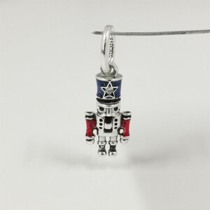 May include: A silver charm shaped like a toy soldier nutcracker. The nutcracker has a blue and silver hat with a star, red accents, and a diamond. The charm is suspended from a silver loop.