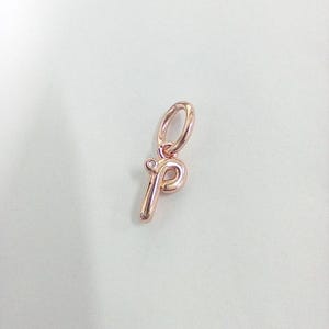 May include: Rose gold charm in the shape of the letter "p". The charm has a small loop at the top for attaching to a bracelet or necklace. A small, clear gem is set at the top of the letter.
