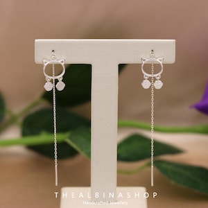 Könnte beinhalten: Ein Paar silberne Ohrhänger mit einem Katzenkopfdesign und kleinen, glitzernden Anhängern, die an der Unterseite hängen. Die Ohrringe werden auf einem weißen Ständer präsentiert.