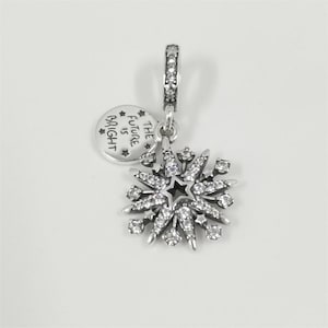 Puede incluir: Un charm de plata con un diseño de copo de nieve y un pequeño charm redondo. El charm de copo de nieve tiene una estrella en el centro y está adornado con cristales transparentes. El charm redondo tiene el texto "THE FUTURE IS BRIGHT".