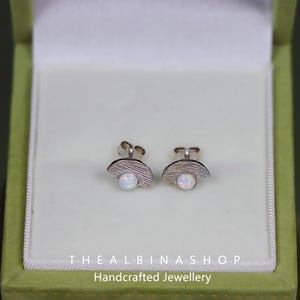 Puede incluir: Un par de pendientes de plata con una piedra de &oacute;palo blanco engastada en forma de media luna. Los pendientes est&aacute;n en una peque&ntilde;a caja verde con el texto "THE ALBINAS SHOP Joyer&iacute;a artesanal".