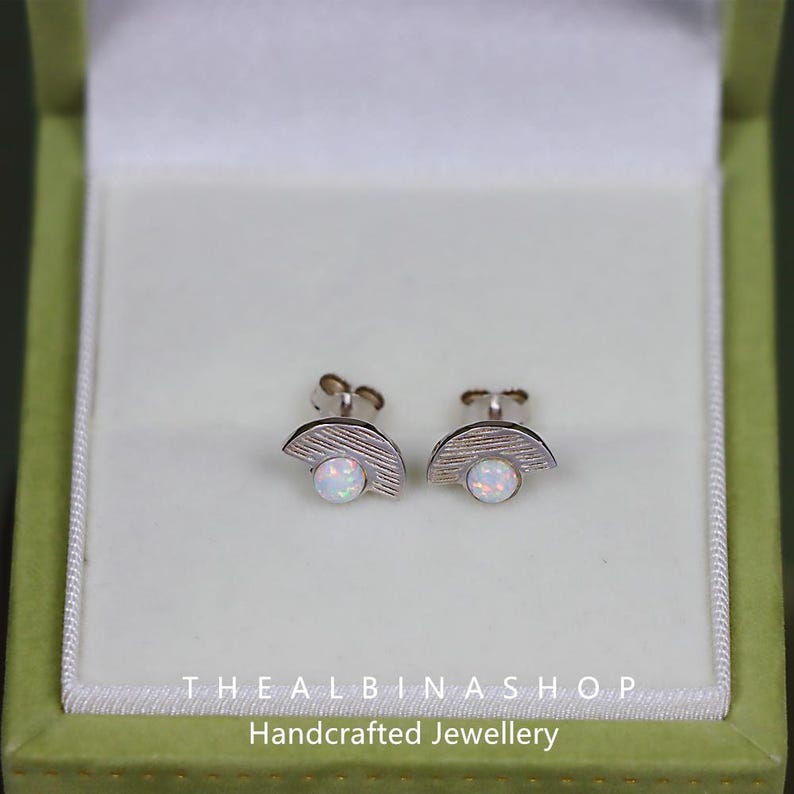 Puede incluir: Un par de pendientes de plata con una piedra de &oacute;palo blanco engastada en forma de media luna. Los pendientes est&aacute;n en una peque&ntilde;a caja verde con el texto "THE ALBINAS SHOP Joyer&iacute;a artesanal".