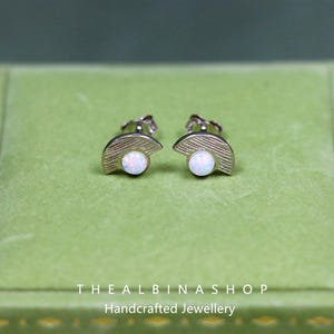 Puede incluir: Un par de pendientes de plata con un dise&ntilde;o de medio c&iacute;rculo y una piedra de &oacute;palo blanco en el centro. Los pendientes se muestran sobre un fondo de terciopelo verde.