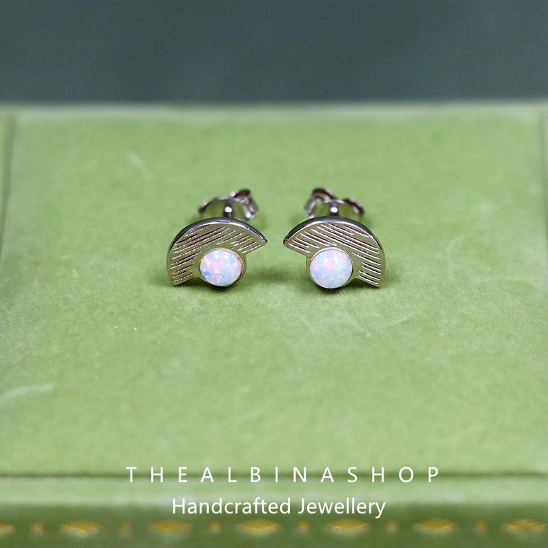 Puede incluir: Un par de pendientes de plata con un dise&ntilde;o de medio c&iacute;rculo y una piedra de &oacute;palo blanco en el centro. Los pendientes se muestran sobre un fondo de terciopelo verde.