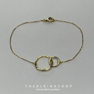 May include: A delicate gold chain bracelet with two interlocking circles. The circles are slightly textured and have a hammered finish. The bracelet is a simple and elegant piece of jewelry.