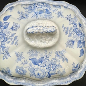 Puede incluir: Una tapa rectangular de cerámica para fuente de servir con base blanca y patrones florales azules. La tapa presenta un borde festoneado y un asa central con forma de hoja. El diseño azul muestra diversas flores y follaje en la superficie.