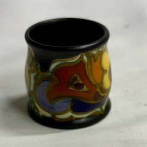 May include: A small, cylindrical ceramic cup with a matte black rim and base. The exterior features a vibrant, abstract pattern of swirling colors including orange, yellow, blue, and brown, set against a cream-colored background.