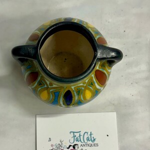 May include: A small ceramic vessel with two black handles, featuring a colorful pattern of blue, yellow, and brown shapes. A square white card is placed below it, displaying the text Fat Cats ANTIQUES with an illustration of a vintage car.
