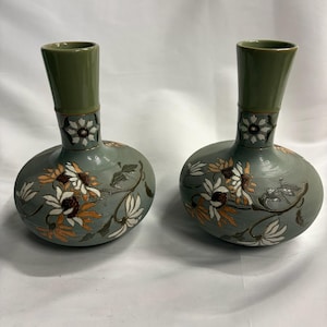 May include: A pair of sage green ceramic vases with a rounded base and a tall, slender neck. The surface features a detailed floral pattern with white and orange petals, dark brown centres, and green leaves, accented by fine horizontal lines.