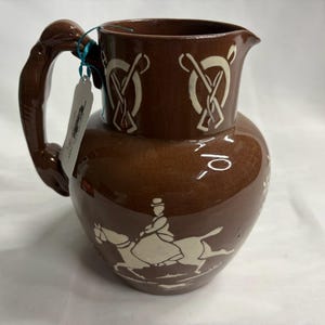 May include: A brown ceramic pitcher featuring a glossy finish and a white graphic print of a person riding a horse. The upper section displays white circular patterns, and a curved handle is attached to the side with a small white paper tag.
