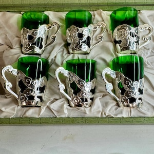 Puede incluir: Un juego de seis tazas de cristal verde esmeralda, cada una dentro de un soporte de metal plateado con motivos en relieve de dragones y nubes, dispuestas en una caja de presentación con platillos de metal plateado a juego debajo.