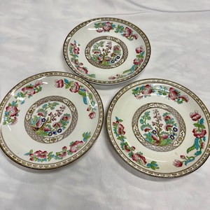 Può includere: Tre piattini rotondi in ceramica con un medaglione centrale raffigurante un motivo ad albero circondato da un bordo geometrico marrone. I bordi sono decorati con un motivo ripetitivo di fiori rosa, foglie verdi e accenti blu su base crema.