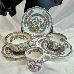 May include: A collection of porcelain tableware featuring two teacups with matching saucers, one small mug, and one decorative plate. Each piece is decorated with a colorful floral pattern of pink flowers and green leaves, accented by gold trim.