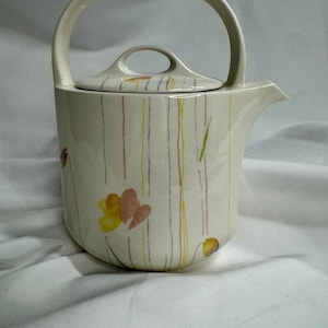 May include: A cream-colored ceramic teapot featuring a tall, arched handle and a matching lid with a small loop handle. The body is decorated with thin, vertical multicolored stripes and soft, painted floral accents in yellow and muted pink tones.