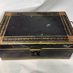 May include: A rectangular black metal storage box with a hinged lid and two side handles. The lid features a decorative gold and orange border pattern with stylized leaf motifs. The box has a central metal latch and shows signs of surface wear.