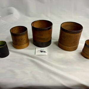Puede incluir: Un conjunto de cinco recipientes de medición cilíndricos de madera de varios tamaños, con bandas metálicas oscuras en los bordes superior e inferior. Los recipientes tienen superficies de madera natural y algunos muestran el texto estampado LITRE.
