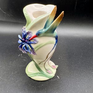 May include: A cream-colored ceramic vase featuring a three-dimensional hummingbird with blue, green, and orange wings and a red beak. The vase is decorated with a sculpted purple iris flower and green stems that wrap around the base and body.