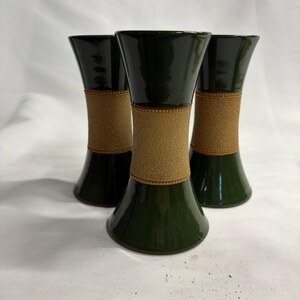 Può includere: Tre vasi a forma di clessidra con una finitura verde scuro lucida sulle sezioni superiore e inferiore, separate da una fascia centrale in materiale beige testurizzato con bordi perlati in alto e in basso, disposti in un gruppo.