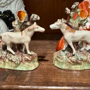 May include: A pair of ceramic horse figurines, each standing on a textured, earth-toned base with a tree-like structure behind the animal. The horses are cream-colored with reddish-brown manes and tails, featuring detailed facial features and hooves.