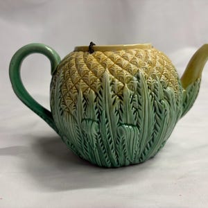 Może przedstawiać: Ceramiczny czajniczek w kształcie ananasa z teksturowanym żółtym korpusem i zielonymi liśćmi u podstawy. Czajniczek posiada zakrzywiony zielony uchwyt po lewej stronie oraz spiczasty zielony dzióbek po prawej stronie, z gładkim otworem u góry.