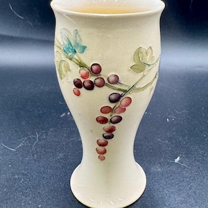 May include: A cream-colored ceramic goblet featuring a hand-painted design of purple grapes and green leaves. The vessel has a flared rim and a tapered base, with a smooth, glossy finish that highlights the detailed botanical illustration on the front.