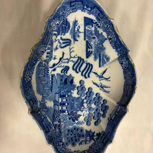 May include: An oval-shaped ceramic dish featuring a blue and white willow pattern. The design includes detailed illustrations of pagodas, a bridge, trees, and two birds in flight. The dish has a scalloped edge and a glossy finish throughout.