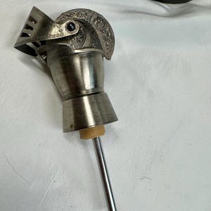 Könnte beinhalten: Ein silberfarbener Flaschenverschluss in Form eines Ritterhelms. Der Helm ist mit detaillierten Prägungen, einem beweglichen Visier und einem kleinen dunklen Akzent versehen. Ein beigefarbener Gummistopfen und ein dünner Metallstab ragen heraus.